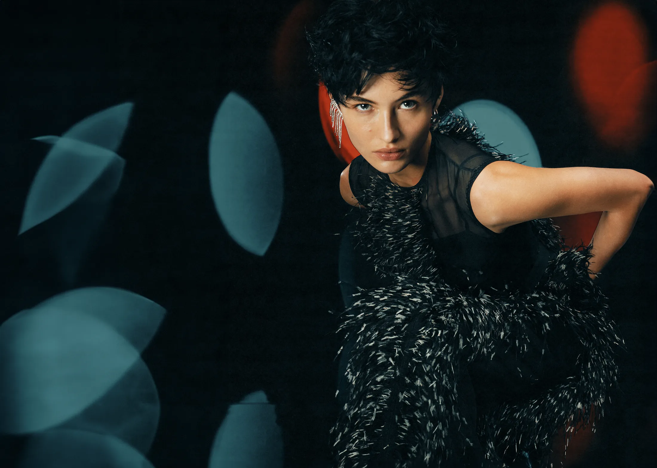A model in a black feathered top against dark teal, one arm raised behind her head.
