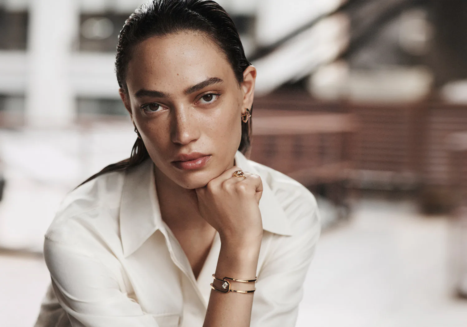 A model in a white shirt wearing gold jewellery, a city behind her.