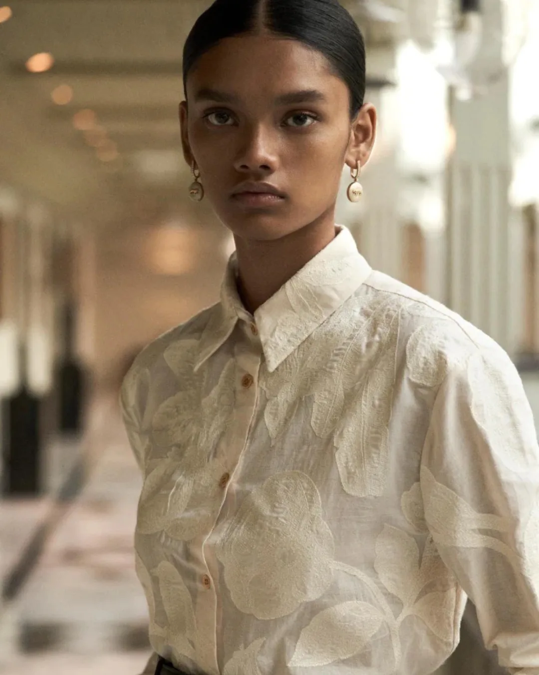 A woman in a cream textured shirt with pearl earrings, a colonnade blurred behind her.