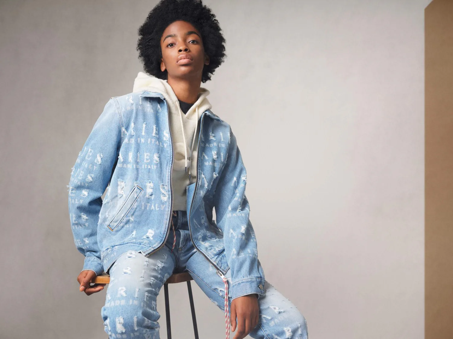 A woman seated on a stool in a pale blue printed denim jacket and matching jeans.