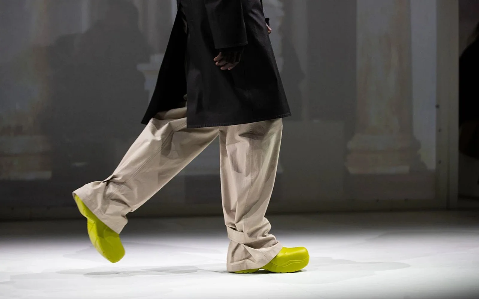 A runway walk seen from the waist down: wide cream trousers and neon yellow shoes.