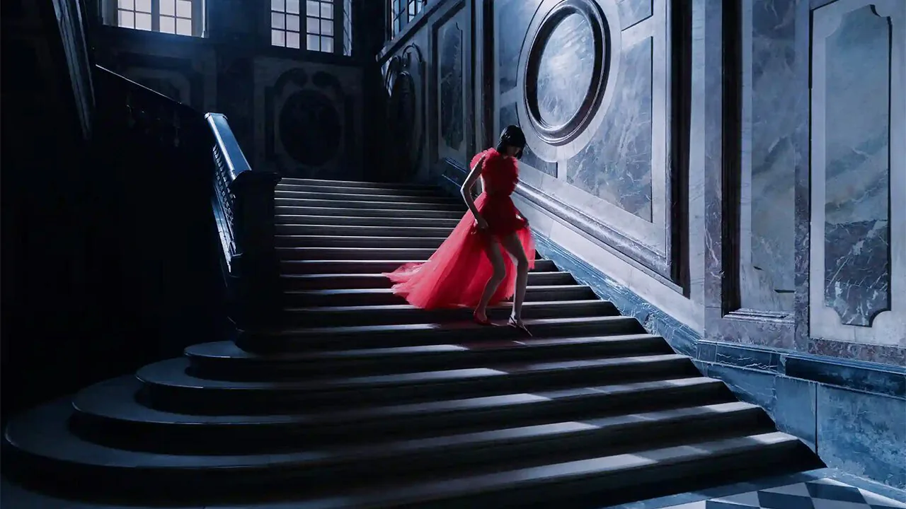 A figure in a red gown on a dark staircase, beneath a large oval mirror.