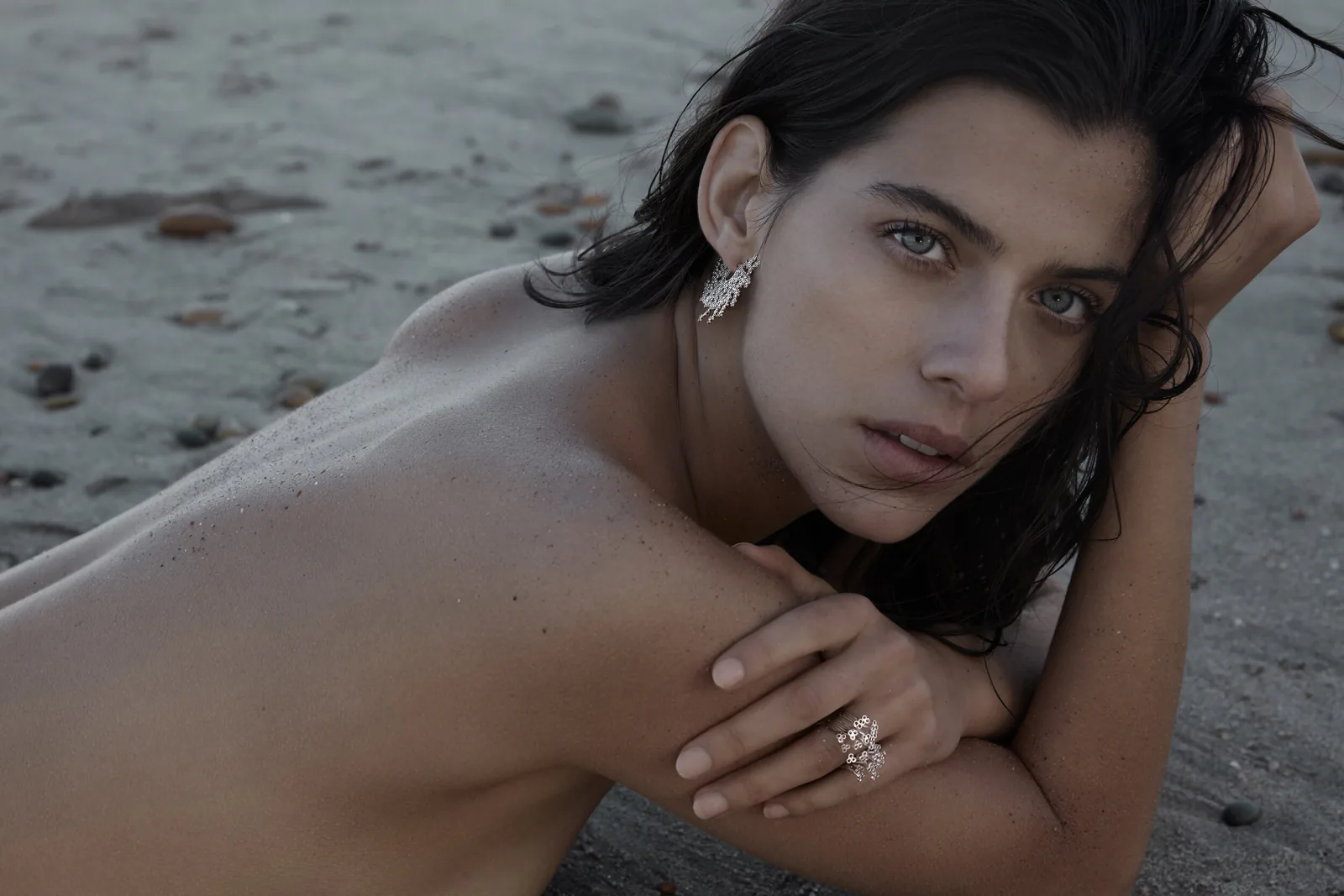 A woman on a pebble beach, one hand on her shoulder, wearing a ring and a drop earring.