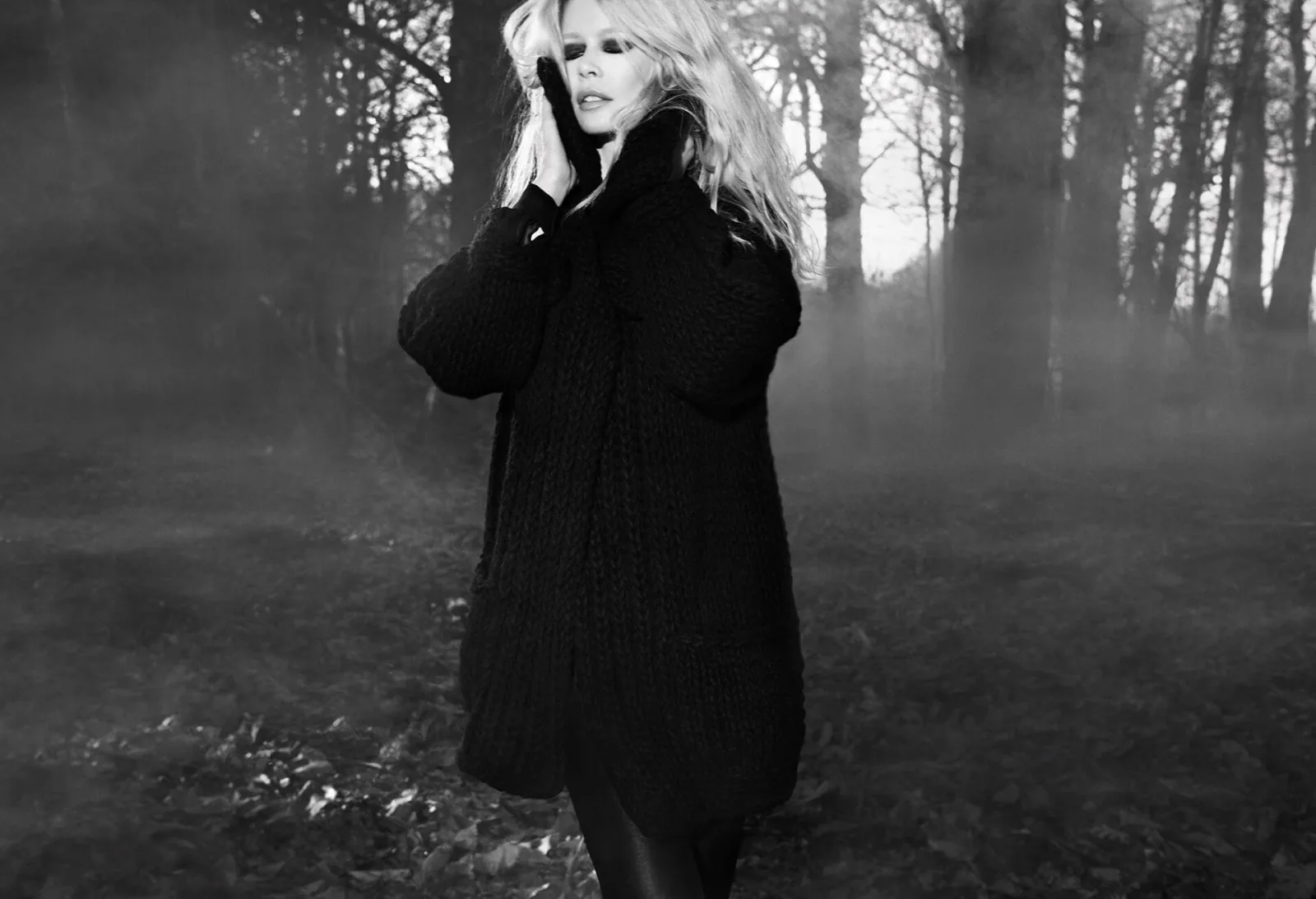 A woman in a dark coat in misty woodland, in black and white.