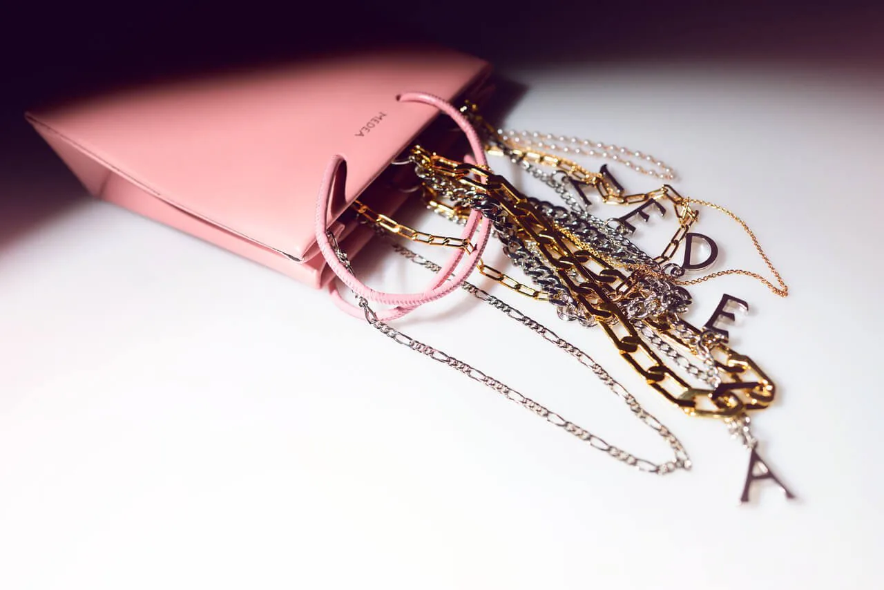 Gold chains and lettered charms spilling from a pink bag on a pale ground.