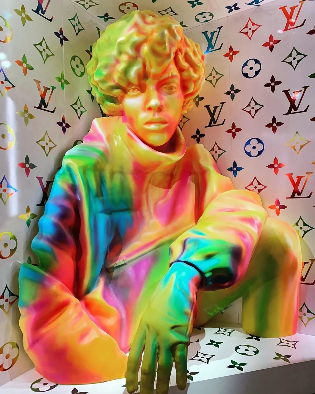 An iridescent rainbow figure seated against a monogrammed wall.