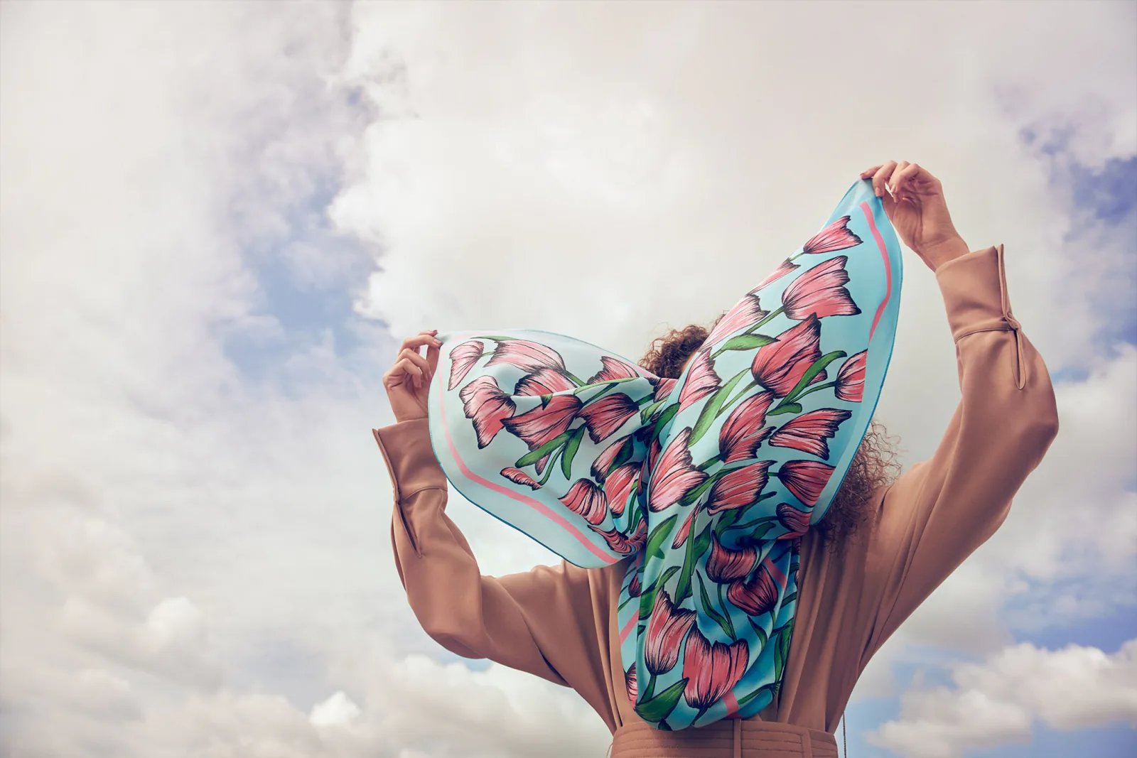 Hands holding a printed silk scarf open against a cloudy sky.