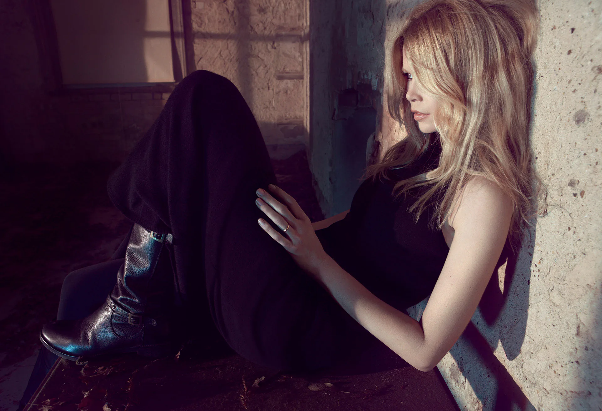 Claudia Schiffer seated against a stone wall in black knitwear and boots.