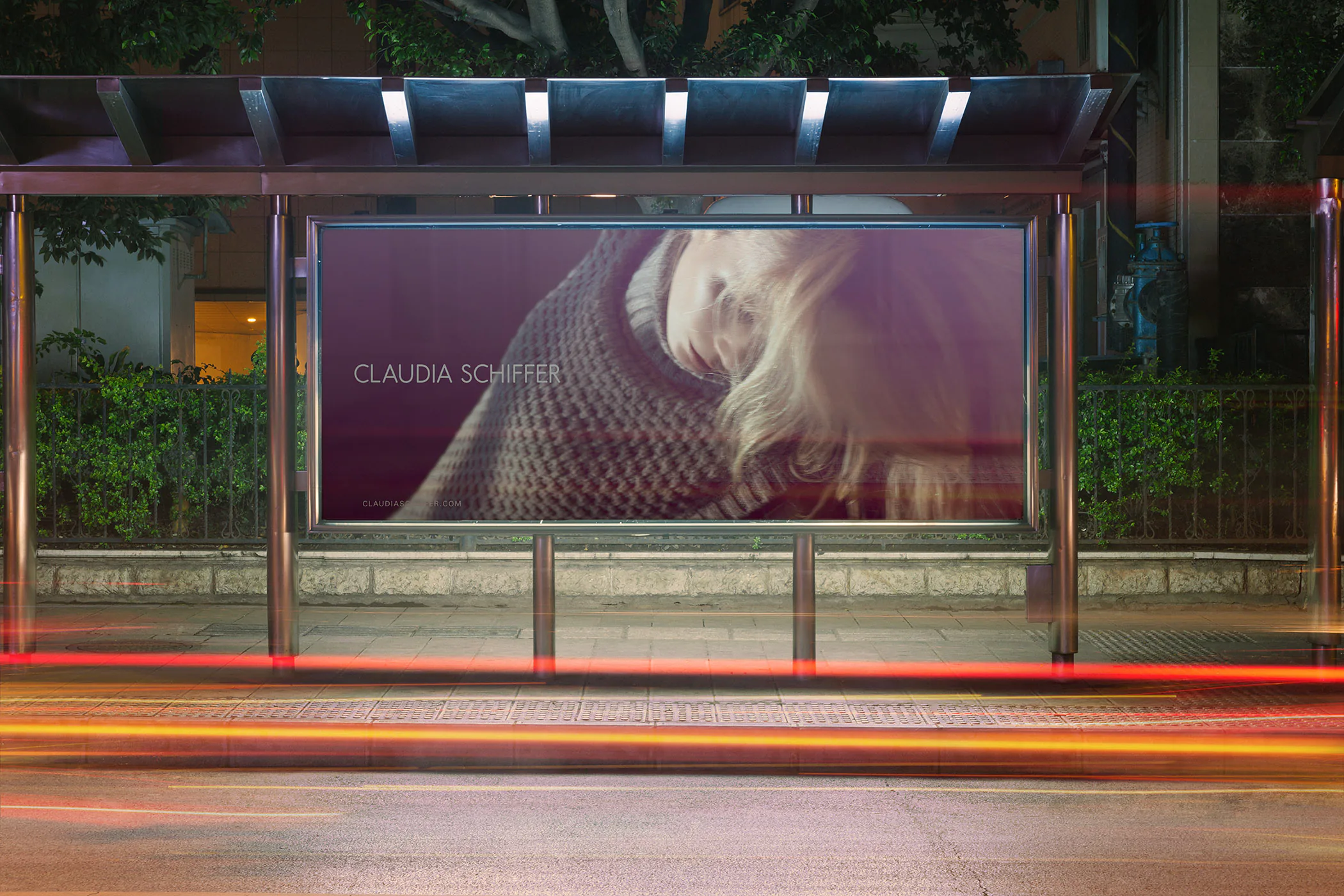 A Claudia Schiffer poster lit in a bus shelter at night, traffic trails passing.