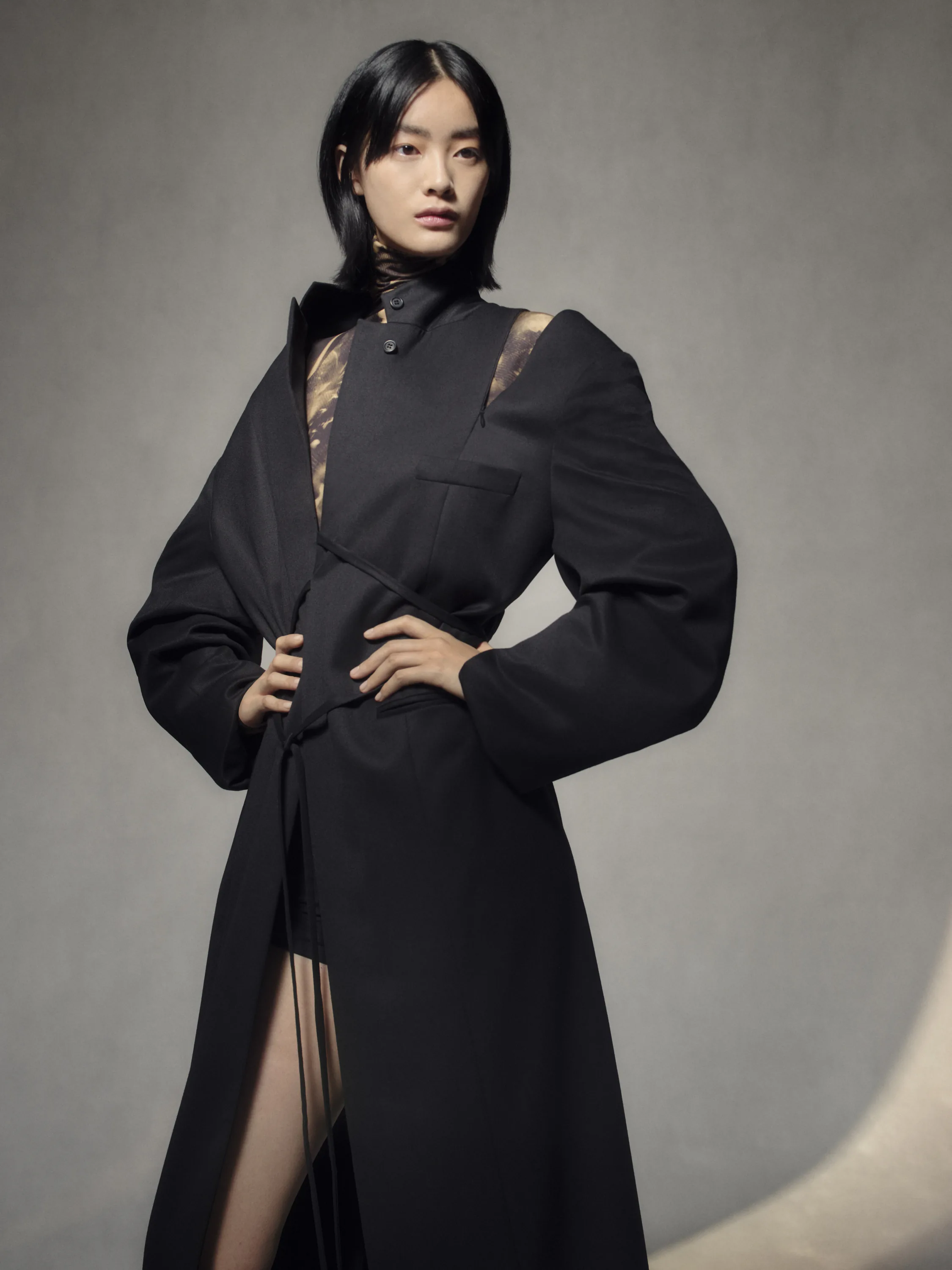 A model in a black belted coat with sheer panels, against a grey backdrop.