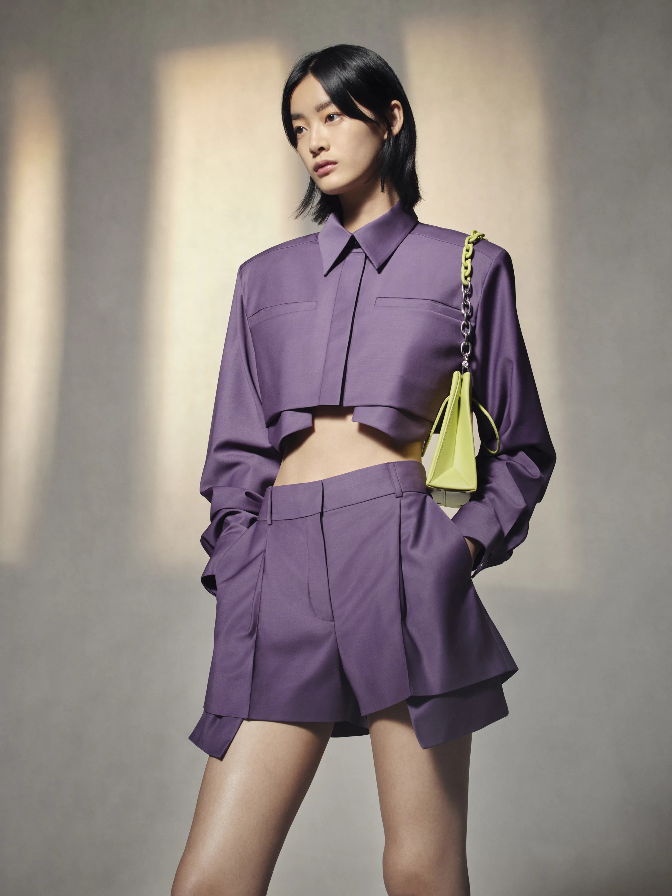 A model in a lilac cropped shirt and shorts with a small yellow bag.