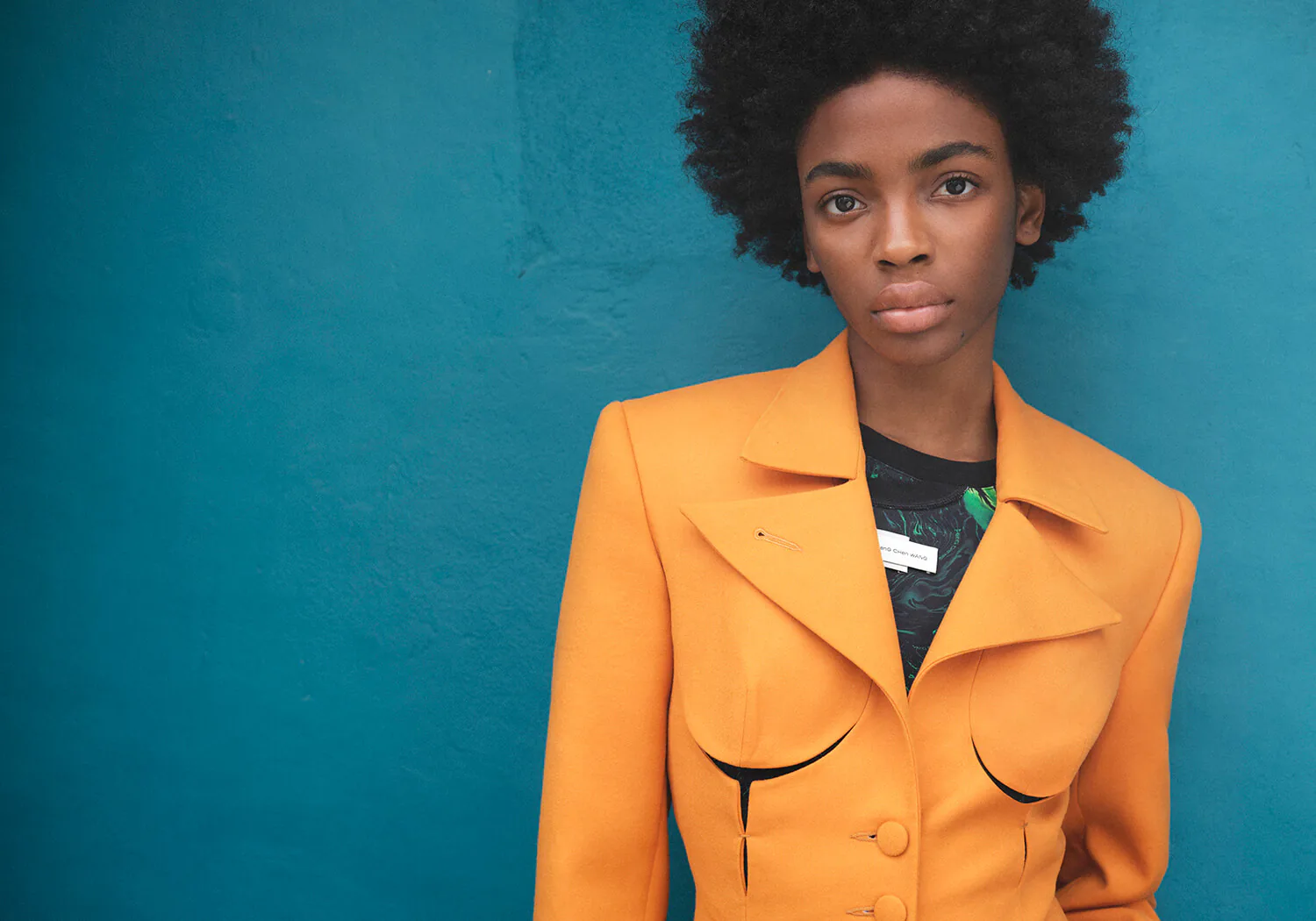 A model in an orange jacket against a teal ground.