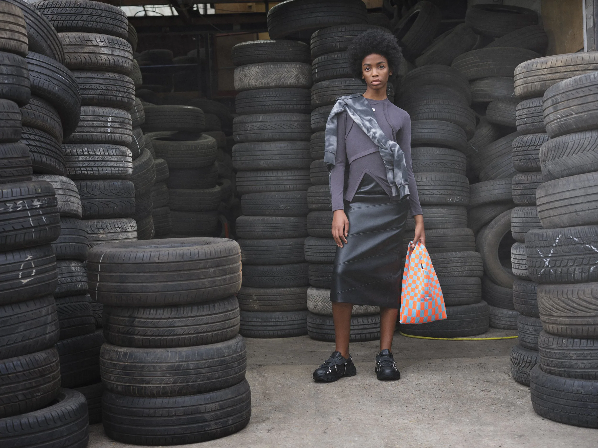 A model in a grey jacket and leather skirt, carrying a patterned bag, in front of walls of stacked tyres.