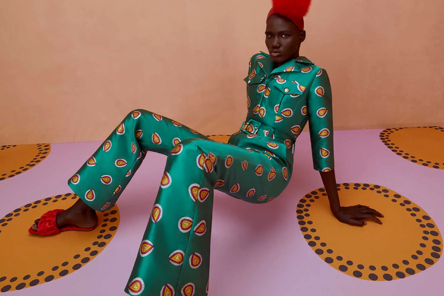 A model in a green and orange printed silk set, seated on a pink and orange floor.
