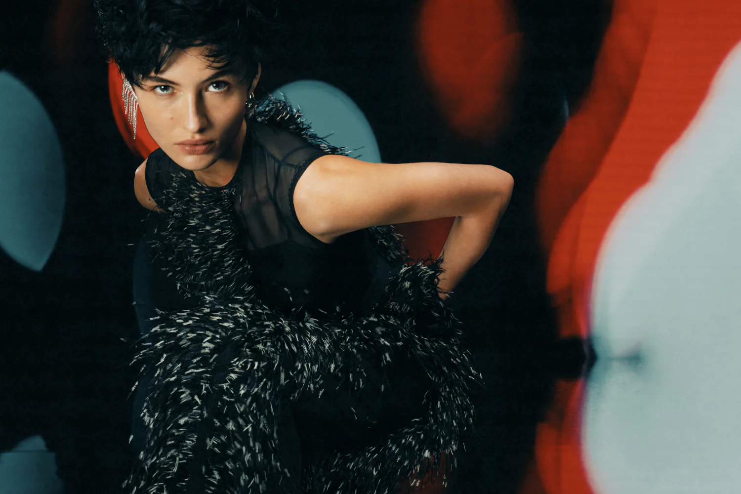 A model in a black feathered dress against a red and teal light wash.