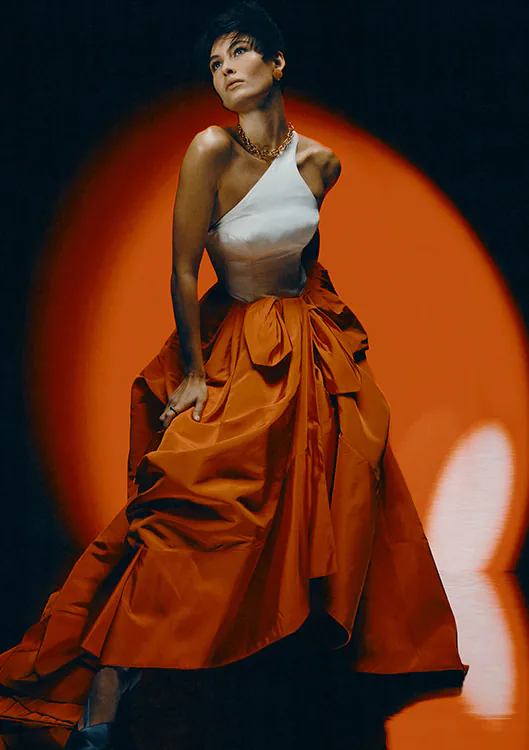 A model in a full orange gown against a dark ground, lit from behind.