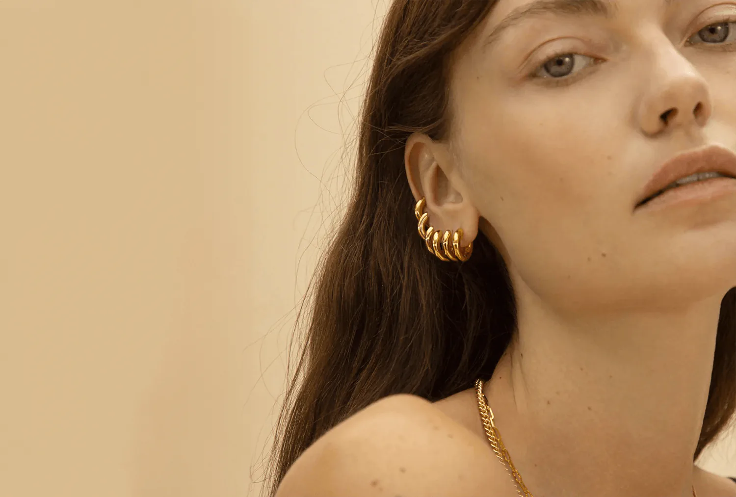 A ribbed gold hoop earring and a fine chain, against a warm sand ground.