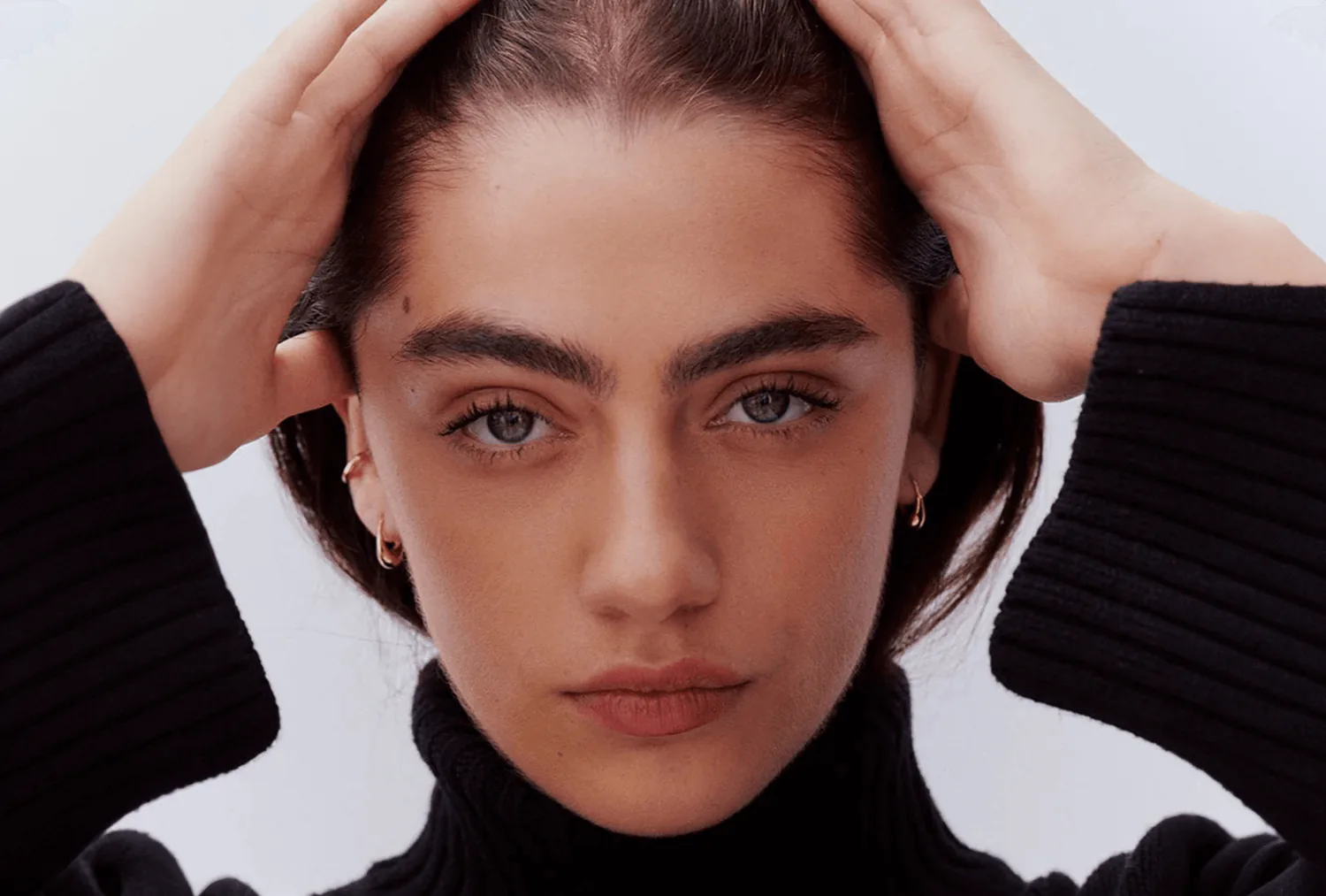 A model looking straight to camera, hands lifted into her hair.