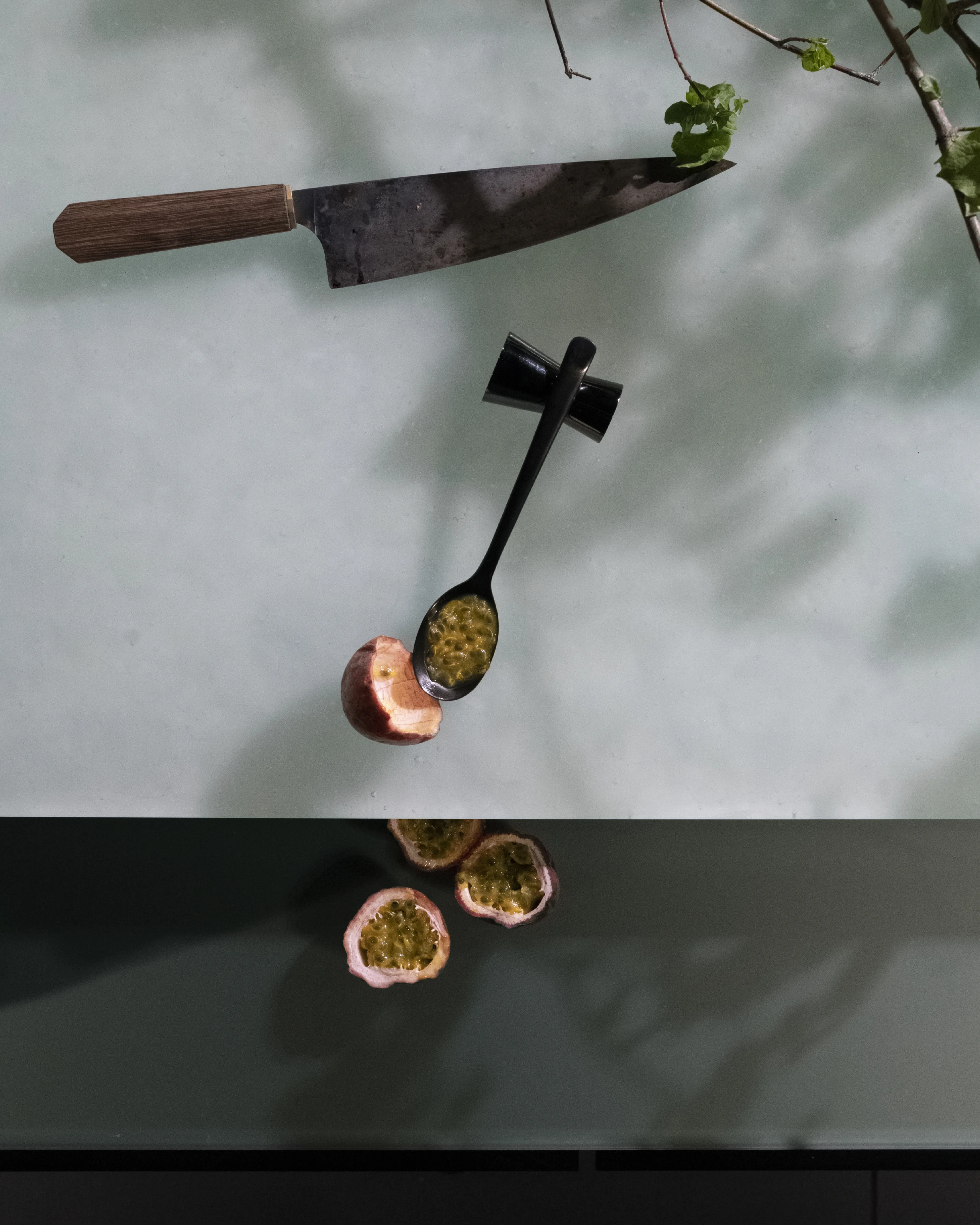 A knife and a spoon of seeds laid on a pale worktop, with halved passion fruit and a trailing stem.