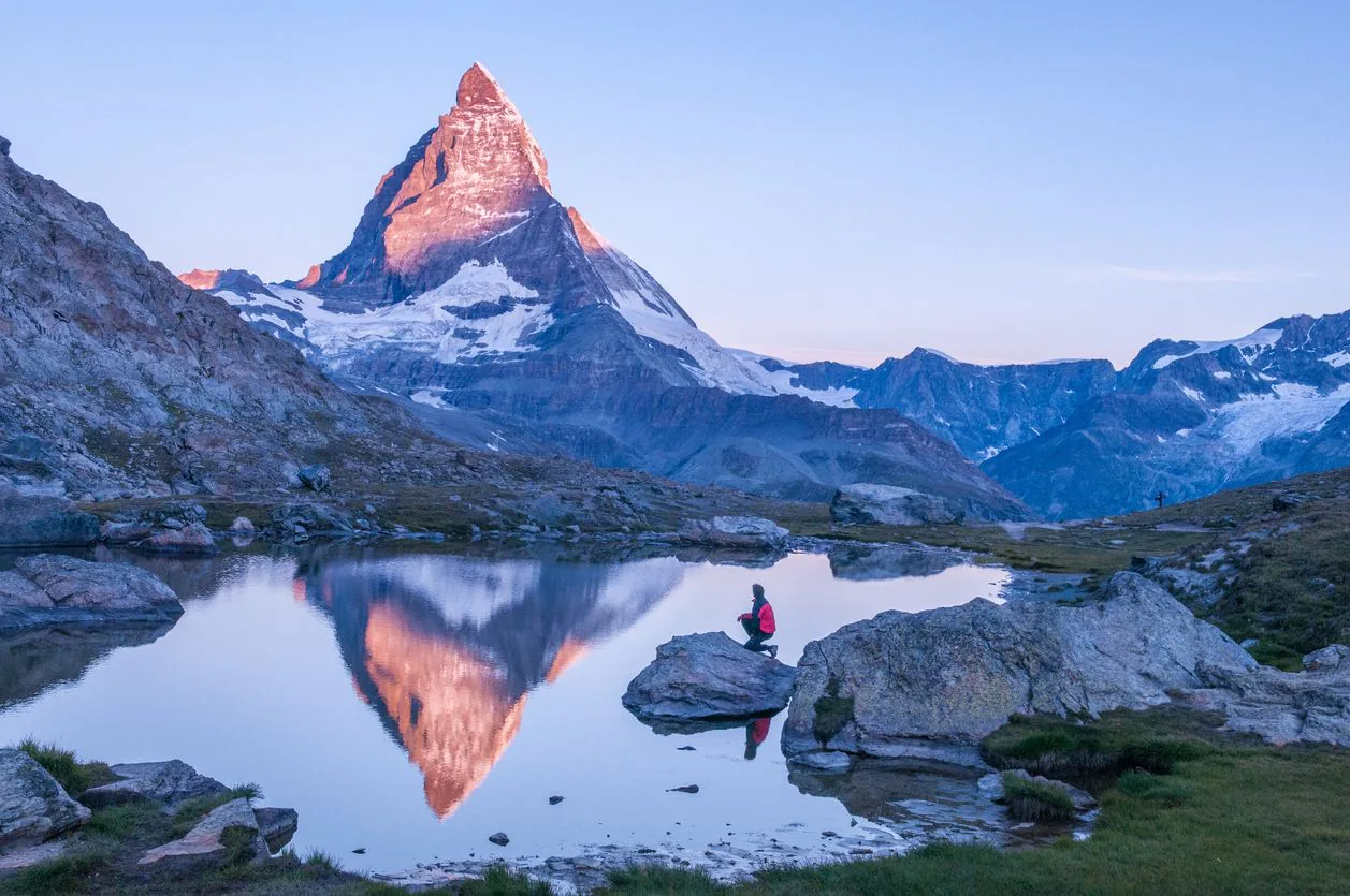 The Matterhorn lit pink at dawn above a lake, with a figure seated on the rocks.
