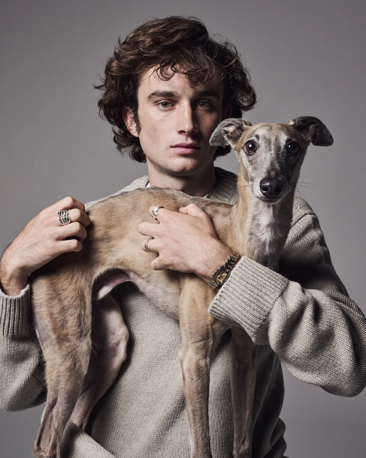 A young man in a cream jumper holding a whippet across his chest.